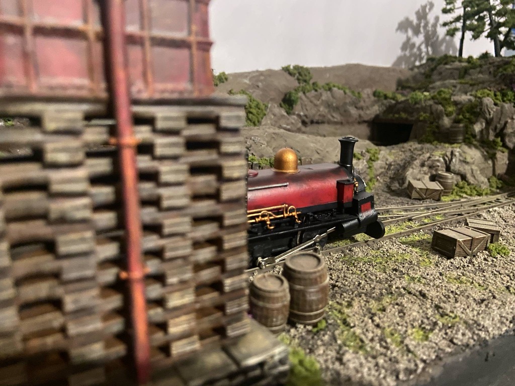 Painted Charles on a narrow gauge layout, seen past a stacked sleeper water tower base with barrels and crates on the ground.