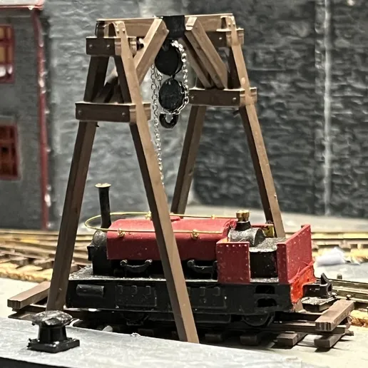 Painted timber A-frame hoist straddling narrow gauge track on a layout, with a chain block lifting over a red and black locomotive.