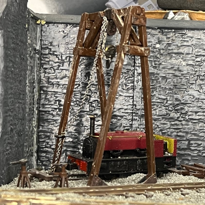 Painted hoist against a stone shed wall, its chain block hanging over a red and black locomotive with two axle jacks beside the track.