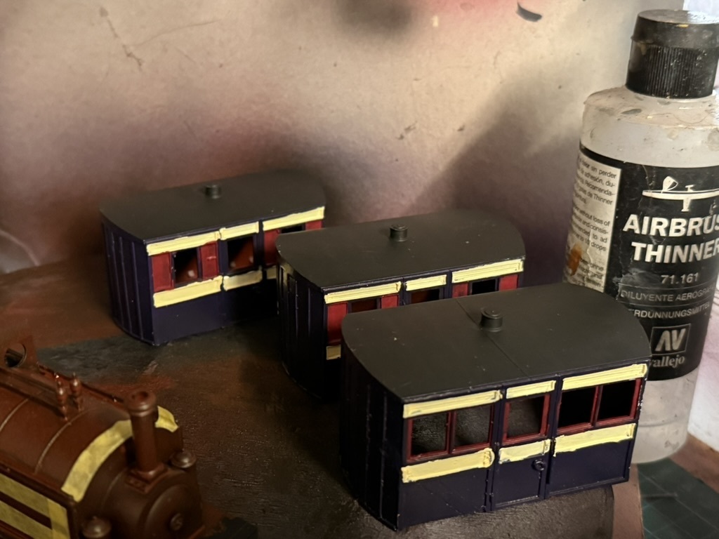Three printed four-wheel coaches part-painted in dark blue with red panels and cream lining, waiting on the workbench beside a bottle of airbrush thinner.